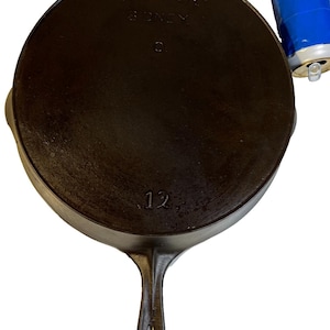 May include: A round, dark brown cast iron skillet with a long handle. The underside features the embossed text WAGNER SIDNEY O. and the number 12. A small, silver-coloured metal can wrapped in blue tape is positioned next to the skillet for scale.