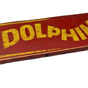 May include: A vintage red wooden skateboard with yellow wheels and the word "DOLPHIN" painted in yellow letters.