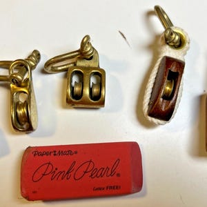 May include: A selection of vintage pulleys in brass and wood, of varying sizes. A red "Pink Pearl" eraser is also present. These nautical items are likely used for rigging or decorative purposes.