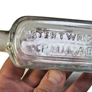 May include: Clear glass bottle with a rectangular body and a narrow neck. The bottle has embossed text that reads "PETER WRIGHT & CO" and "PHILAD". The bottle is held in a person's hand.