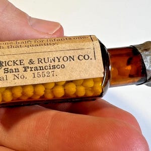 May include: An antique amber glass bottle containing small yellow pellets. The bottle features a paper label with the text "BOERICKE & RUNYON CO. San Francisco" and "Serial No. 15527." The bottle is sealed with a metal cap.