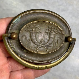 Puede incluir: Tirador de puerta de armario de latón antiguo con una placa ovalada que presenta un diseño en relieve de dos urnas con acentos florales. La placa tiene el número "BR148" grabado en la parte inferior.