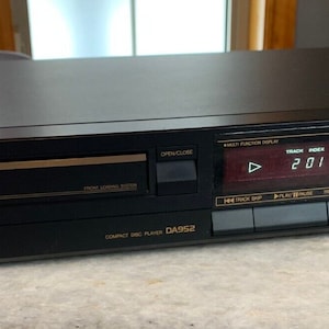 Könnte beinhalten: Ein schwarzer Scott CD-Player mit einem digitalen Display, das "2:01" anzeigt. Der Player hat eine silberne Frontplatte mit goldenen Beschriftungen und Knöpfen. Der Player ist mit "Compact Disc Player DAS92" beschriftet.