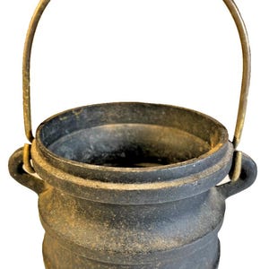 May include: A small, dark gray cast iron cauldron with a rounded body, three short legs, and two side handles. A thin, arched metal handle is attached to the side loops, extending upward above the open top of the vessel.