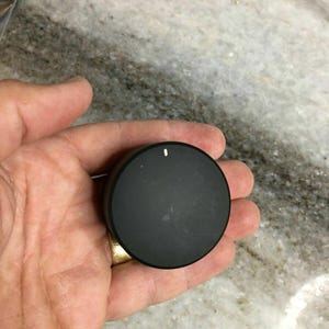 Puede incluir: Un mando negro y circular con una línea indicadora blanca. El mando se sostiene en una mano sobre una superficie similar al mármol. El mando es probablemente un control para un electrodoméstico o dispositivo electrónico.