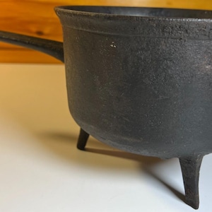 May include: A black cast iron pot with a textured surface, featuring a single long handle extending from the side and three short, sturdy legs supporting the base. The pot has a wide, open circular top and a deep, rounded basin structure.