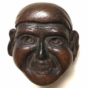 May include: A dark brown wooden carving of a smiling face with a wide mouth and large ears. The face is carved in a realistic style and has a detailed expression.