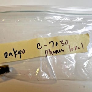 Puede incluir: Un pequeño componente electrónico negro dentro de una bolsa de plástico transparente. Un trozo de cinta adhesiva beige con texto manuscrito que dice "Onkyo C-7030 Phones level" está adherido a la bolsa. El componente parece ser una pequeña pieza de audio.