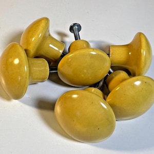 May include: Seven yellow oval cabinet knobs with metal screws, arranged on a white surface. The knobs have a glossy finish and are a light yellow colour. Each knob is designed for easy installation.