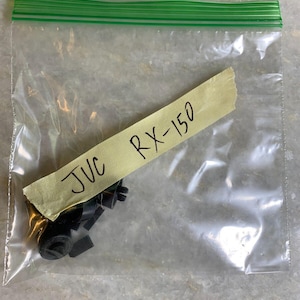 Puede incluir: Una bolsa de plástico transparente con varias piezas pequeñas de plástico negro. Una nota adhesiva amarilla pegada a la bolsa dice "JVC RX-150".