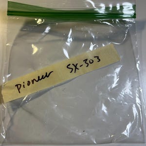 Op de afbeelding: Een doorzichtige, hersluitbare plastic opbergzak met een groene ritssluiting aan de bovenkant. Een rechthoekig stuk beige afplaktape is op de voorkant van de zak geplakt, met daarop de handgeschreven tekst Pioneer SX-303 in zwarte inkt.