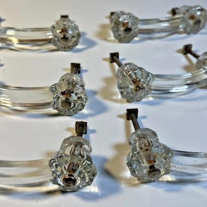 May include: Six clear glass cabinet pulls. Each pull has a curved, clear glass handle with decorative, faceted ends and a metal mounting post. These pulls are designed for easy installation and add a touch of vintage charm.