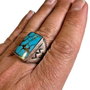May include: A silver ring featuring a rectangular turquoise stone with dark brown matrix veining. The metal band has a wide, thick profile with stamped geometric markings along the side, showcasing a polished yet textured finish on the silver surface.