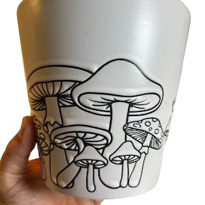 Puede incluir: Una maceta de cerámica blanca con un diseño de contorno negro de setas. La maceta tiene una forma ligeramente cónica y un acabado liso y mate. El diseño de setas es detallado y cubre una parte de la superficie de la maceta.