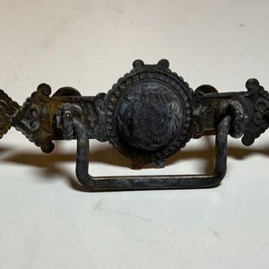 Könnte beinhalten: Drei antike Schubladengriffe aus Bronze. Jeder Griff hat ein zentrales kreisförmiges Design mit aufwendigen Details, flankiert von dekorativen Elementen und einem rechteckigen Griff. Die Griffe haben eine dunkle, verwitterte Oberfläche.
