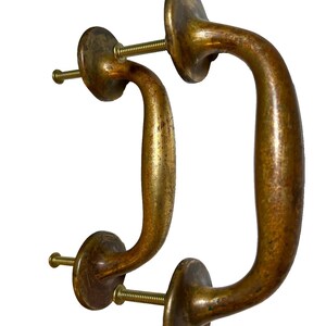 May include: Two antique brass door handles with a curved design. Each handle has a circular base and a curved grip, connected by a threaded screw. The brass has a warm, aged patina, suggesting vintage craftsmanship. The handles are designed for easy installation.
