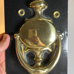 May include: A Classic Solid Brass door knocker by Stanley. The brass knocker has an oval center with a curved handle and a gold finish. The packaging includes the text "Door Knocker" and "LifeSpan".
