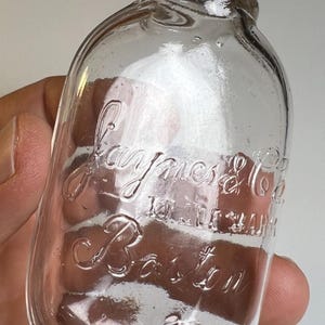 May include: A clear glass bottle with a silver-coloured screw-top lid. The glass features embossed lettering that reads Jaynes & Co. 101 Milk St. Boston. The bottle has a rounded body and a narrow neck, with a small embossed number 2 visible near the base.