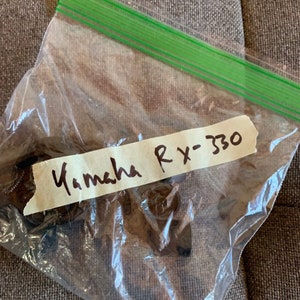Puede incluir: Una bolsa de plástico transparente con cierre de cremallera verde que contiene piezas metálicas marrones. Una etiqueta blanca con escritura negra dice "Yamaha RX-330".