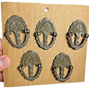 May include: Five antique metal drawer pulls with an ornate design. Each pull has a central floral motif and a decorative border. The pulls are attached to cardboard, suggesting they are for sale or display. The pulls are made of metal.
