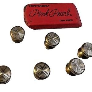 May include: A red Paper Mate Pink Pearl eraser with the text "Pink Pearl" and "Latex FREE!" is positioned above six silver-toned knobs with a spiral design. The knobs are arranged around the eraser.