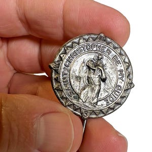 Könnte beinhalten: Ein silberfarbener St. Christopherus-Ansteckpin mit der Aufschrift "SAINT CHRISTOPHER BE MY GUIDE". Der Pin zeigt den Heiligen Christophorus, der ein Kind trägt. Der runde Pin hat eine spitze Nadel auf der Rückseite.