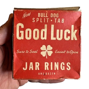 Op de afbeelding: Een rode doos met "Good Luck" potringen, vastgehouden in een hand. De doos heeft witte tekst en een klaverblad-afbeelding. Tekst bevat "New", "Bull Dog Split Tab", "Sure to Seal", "Easiest to Open" en "One Dozen."