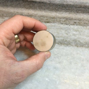 Puede incluir: Un recipiente redondo de color plateado con una tapa beige. La tapa tiene una superficie ligeramente texturizada.