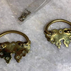 Peut inclure: Deux poignées de tiroir en laiton ornées d'un motif floral décoratif. Les poignées ont une poignée incurvée et une plaque arrière décorative.