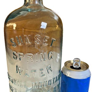Puede incluir: Una botella de vidrio vintage con texto en relieve que dice "SUN SET SPRING WATER" y "ESKILL MOUNTAIN, Haines Falls, NY". Una lata azul y plateada está al lado de la botella. La botella mide aproximadamente 25 cm de altura.