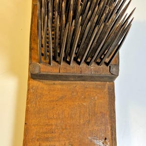Op de afbeelding: Een vintage vlashekkel met een rechthoekige houten basis in een verweerde bruine afwerking en een dichte reeks lange, scherpe, rechtopstaande metalen pinnen die door een metalen frame worden vastgehouden. De houten plank heeft inkepingen.