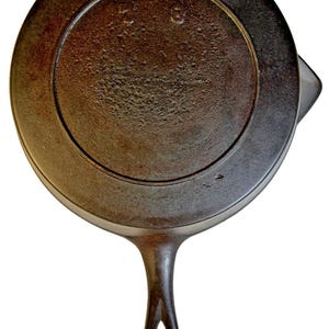 Puede incluir: Sartén de hierro fundido, de color marrón oscuro, con mango largo y orificio para colgar. La superficie de cocción circular tiene los números "7" y "8" grabados. La sartén, de unos 20 cm de diámetro, muestra signos de uso.