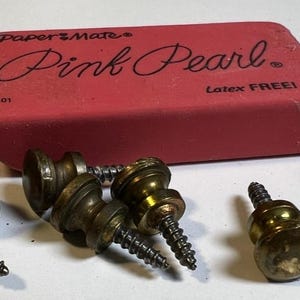 Può includere: Una collezione di viti color ottone con teste decorative, sparse attorno a una gomma rosa rettangolare. La gomma è etichettata "Paper Mate Pink Pearl" con il testo "Latex FREE!"