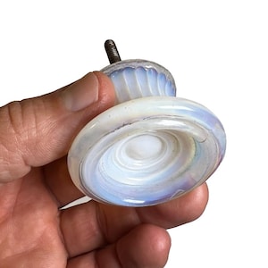 May include: A round, translucent white glass cabinet knob with an iridescent, opalescent finish. The knob features a fluted, ribbed neck and a flat, circular face with concentric ring detailing. A threaded metal screw extends from the back.