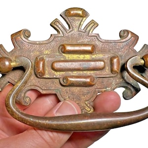 Può includere: Maniglia per cassetto in bronzo antico con dettagli ornati. La maniglia presenta una placca posteriore decorativa con un design rettangolare centrale e una maniglia curva. L'hardware è tenuto in mano su sfondo bianco.