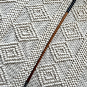 Könnte beinhalten: Ein Vintage-Golfschläger mit Holzschaft und schwarzem Griff. Der Schlägerkopf ist silberfarben und abgewinkelt. Der Schläger liegt auf einem strukturierten, cremefarbenen Teppich mit geometrischem Muster.