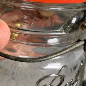 May include: Close-up of a clear glass jar with a red lid and metal wire closure. The jar has the embossed letters "RIV" on the side. The jar is partially filled and appears to be vintage.