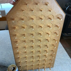 Op de afbeelding: Een houten pegboard-spel met een lichtbruine afwerking en rijen houten pinnen. Het bord heeft de vorm van een huis.