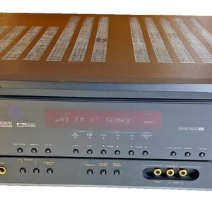 May include: A black Denon audio receiver with a digital display showing radio frequency information. The receiver has multiple buttons, knobs, and input jacks. The top of the receiver has a series of horizontal vents.