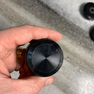 Puede incluir: Un pomo redondo negro con un centro ligeramente elevado. El pomo es liso y tiene un acabado brillante.