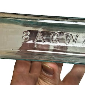 May include: A clear, rectangular glass bottle with the embossed word "SAGWA". The bottle has a rounded neck and a flat, rectangular body. The glass has a slight blue-green tint.