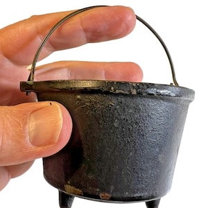 May include: A small, black cast iron cauldron with three short legs and a wire handle. The cauldron has a textured surface and is held in a hand against a white background. This miniature cauldron is suitable for decorative purposes.