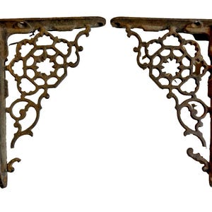 May include: Two antique, cast iron shelf brackets. The brackets are a dark, rusty brown color with ornate, openwork designs. Each bracket has a right-angle shape with mounting holes for easy installation. The decorative elements include floral and scroll patterns.