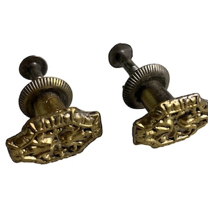 Könnte beinhalten: Zwei goldfarbene Schrankknöpfe mit verzierten Designs und einer silbernen Schraube. Die Knöpfe haben eine quadratische Form mit abgerundeten Kanten und aufwendigen Details.