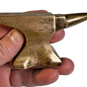 Könnte beinhalten: Ein kleiner Amboss aus Messing, gehalten in einer Hand. Der Amboss hat einen rechteckigen Körper und ein spitzes Horn. Das Metall hat einen goldenen Farbton und weist Gebrauchsspuren auf. Es ist wahrscheinlich ein Dekorationsartikel.