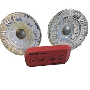 May include: Two round, clear glass cabinet knobs with a textured, scalloped edge and a central metallic silver screw head, positioned behind a rectangular pink Paper Mate Pink Pearl eraser with black text that reads Latex FREE.