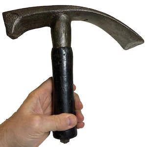 Puede incluir: Un pico de metal oscuro con una hoja curva y un mango de madera. El pico se sostiene en una mano, destacando su diseño. La herramienta parece usada, posiblemente una antigüedad. El mango es oscuro y el metal tiene un aspecto desgastado.