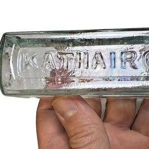 May include: A rectangular, clear glass bottle with the embossed text "KATHAIRON". The bottle has a rounded neck and a rectangular body. The bottle is held in a hand.