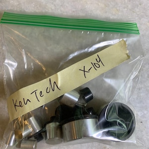 Puede incluir: Una bolsa de plástico transparente con cierre de cremallera verde que contiene varios componentes metálicos y negros. Una etiqueta amarillenta dice "Ken Tech X-104" con rotulador negro. La bolsa está sobre una superficie gris claro.
