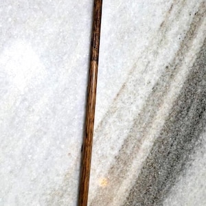 Könnte beinhalten: Ein antiker Golfschläger mit einem dunklen Holzschaft und einem braunen Schlägerkopf. Der Griff ist mit einem dunklen, strukturierten Material umwickelt. Der Schläger steht vor einem Marmorhintergrund.